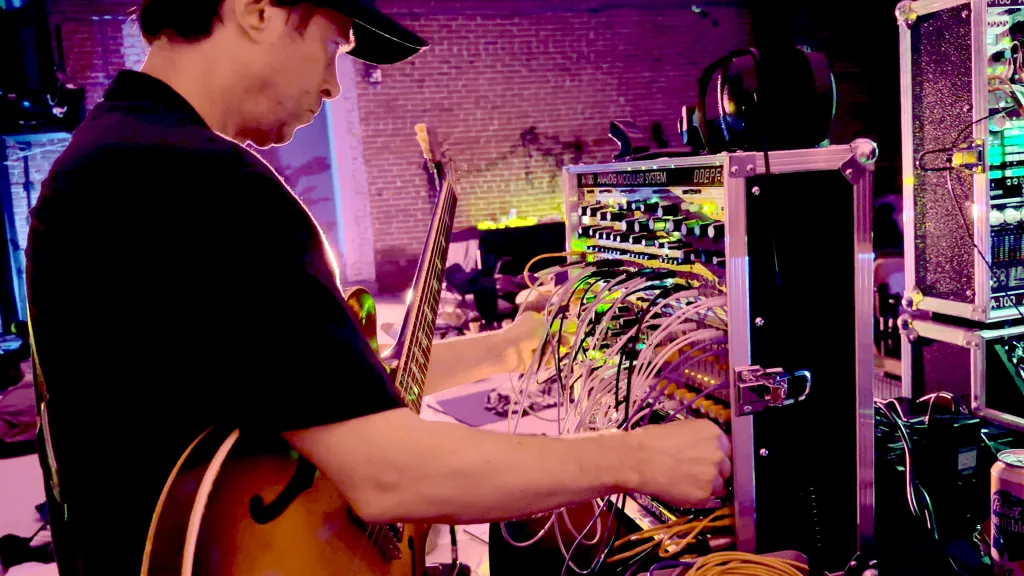 Musician playing a hollow-body guitar while operating a modular synthesizer with many patch cables.
