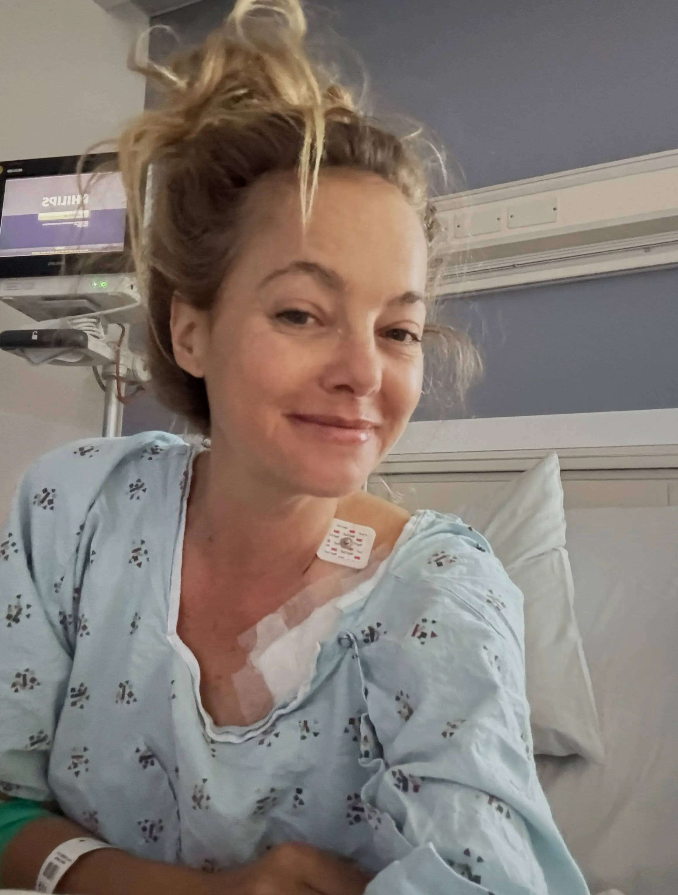 Bijou Phillips in a hospital gown with a medical patch on her chest, smiling at the camera.