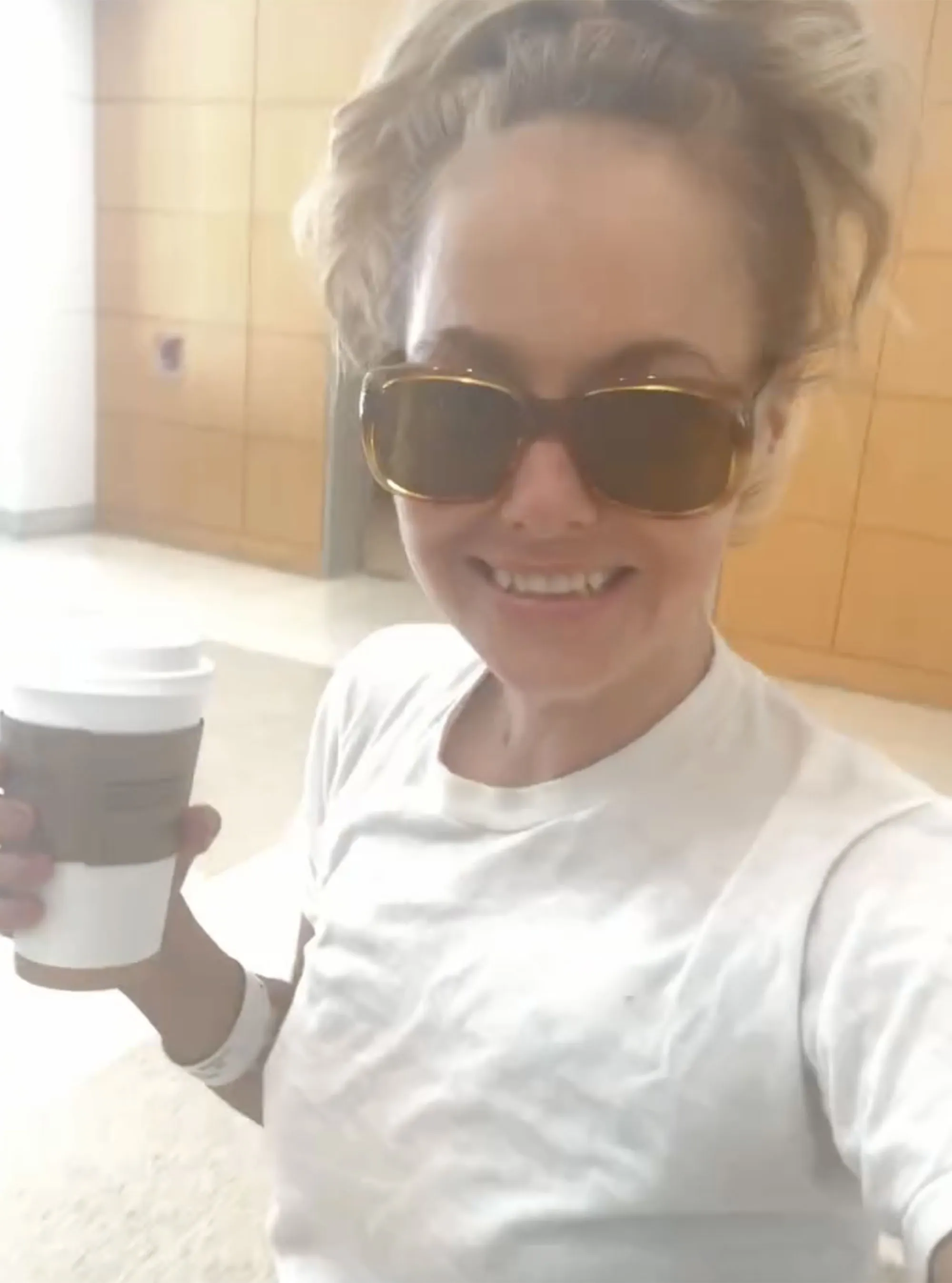 Bijou Phillips, in sunglasses and a white t-shirt, smiling and holding a coffee cup.
