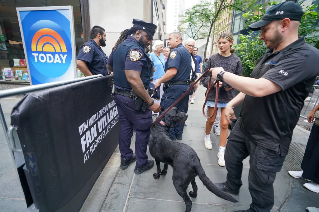 Security checkpoint with NYPD officers, a K-9 unit, and a black dog outside the NBC TODAY show taping at Rockefeller Plaza.