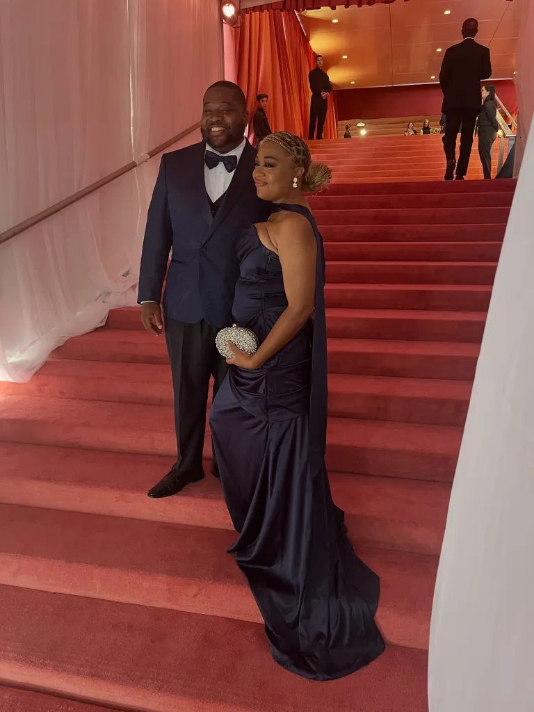 A couple in formal attire posing on a red carpeted staircase.