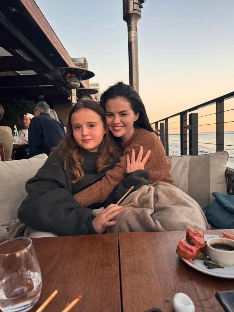 Selena Gomez and her younger sister sit at an outdoor table with a plate of food, a drink, and chopsticks, overlooking the ocean at dusk.