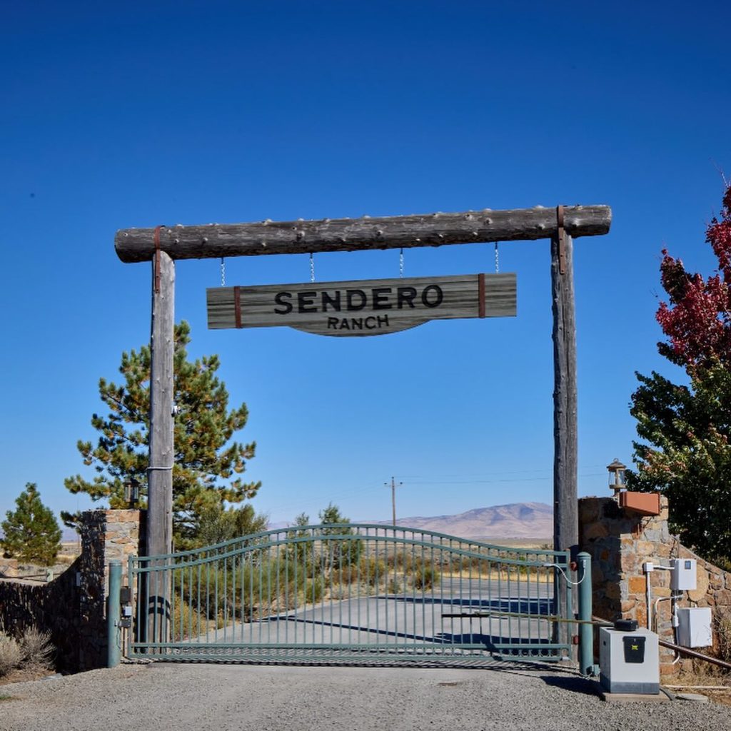 A wooden sign for "Sendero Ranch" hangs above a green metal gate, with a road extending into a desert landscape with mountains in the background.