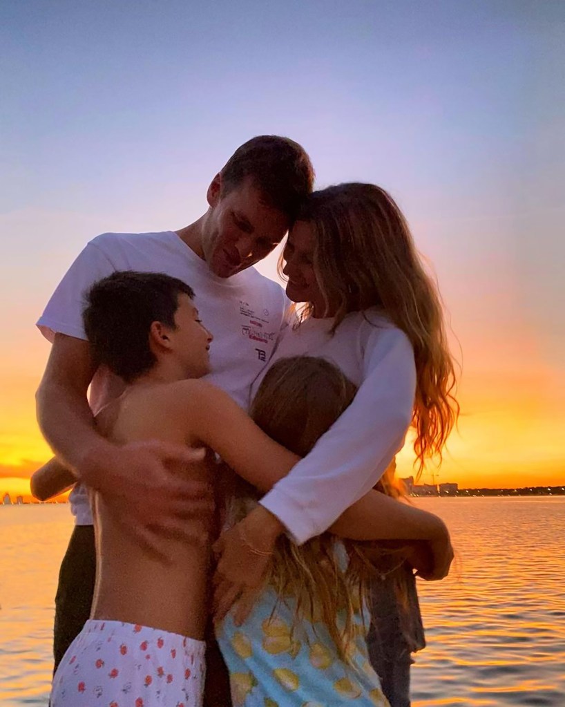A family of four, including two adults and two children, embraces against a sunset over water.