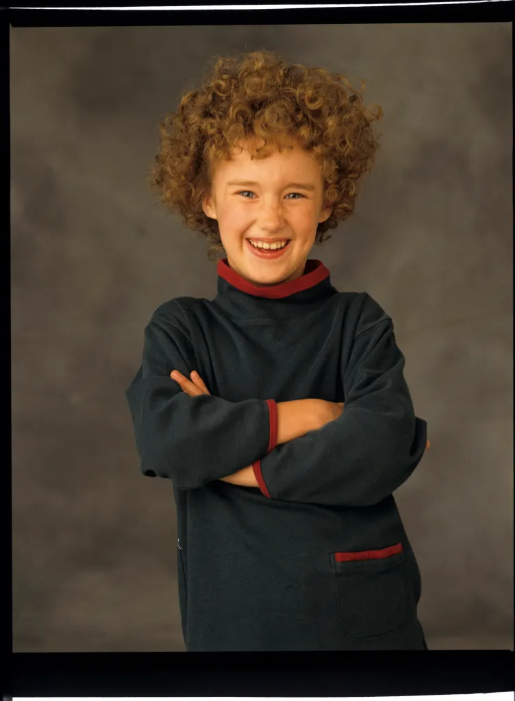 Ashley Johnson, a young girl with curly red hair and freckles, wearing a dark green long-sleeved shirt with red trim, smiling with her arms crossed.