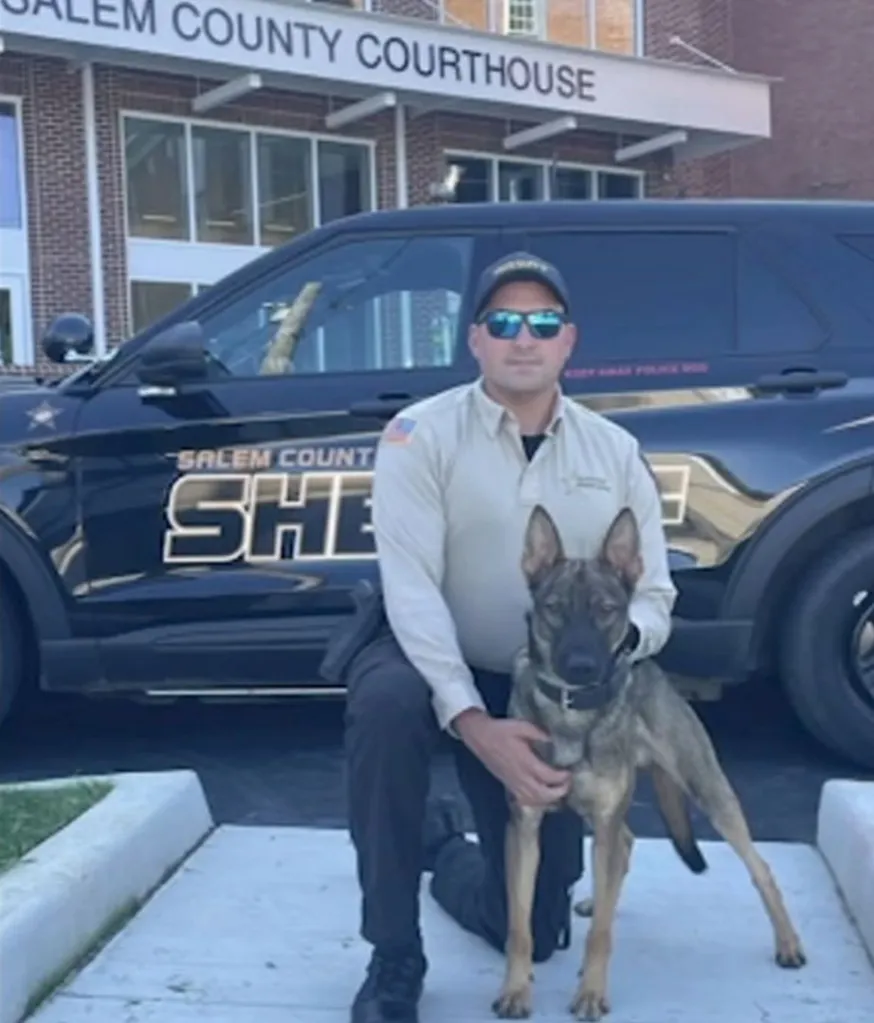 Sgt. Cody Henderson kneeling beside K9 Rip, with a Salem County Sheriff vehicle and courthouse in the background.
