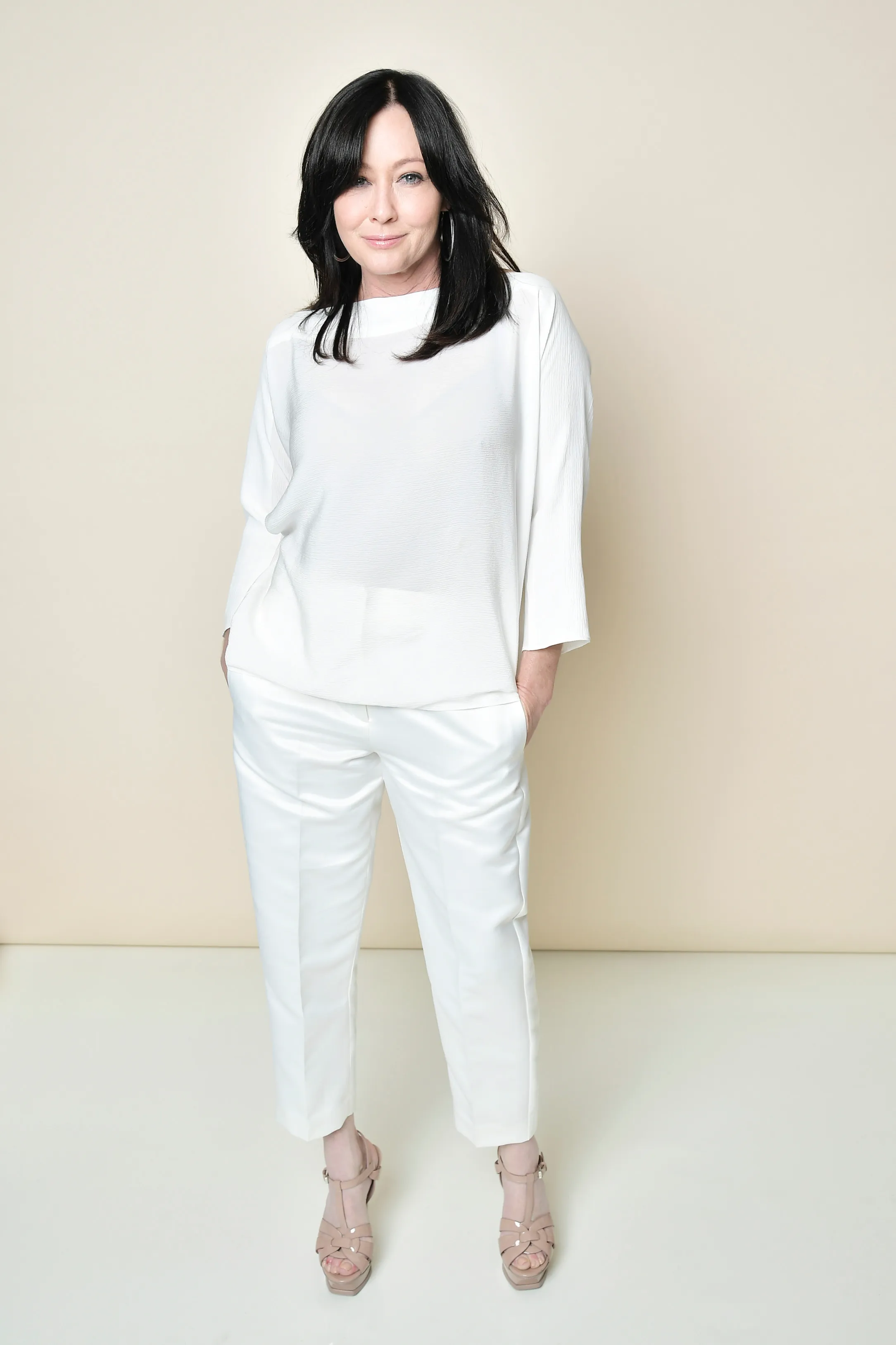Shannen Doherty posing for a portrait in a white top and pants.