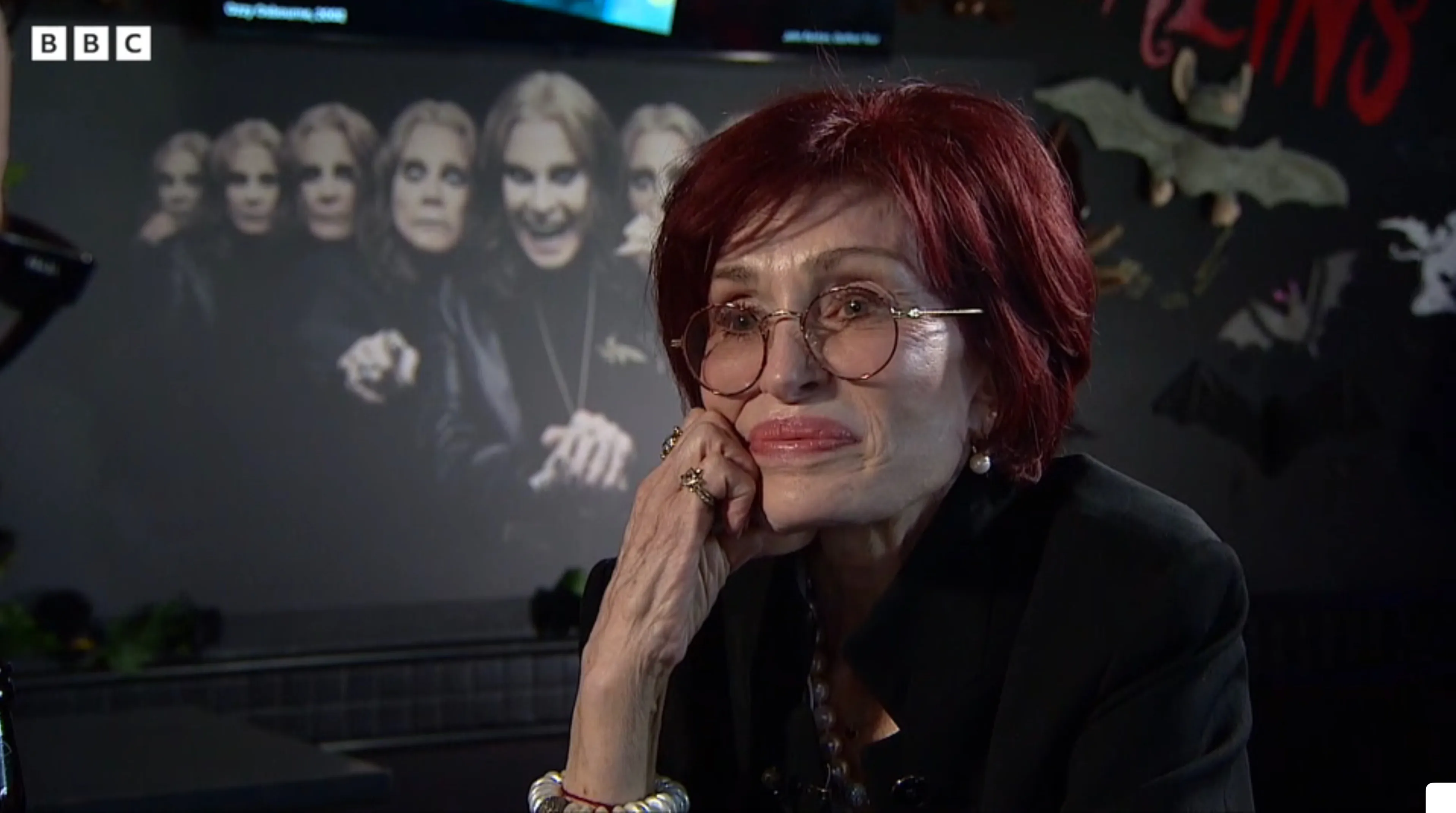 Sharon Osbourne, with red hair and glasses, reflects on her husband Ozzy with his images in the background.