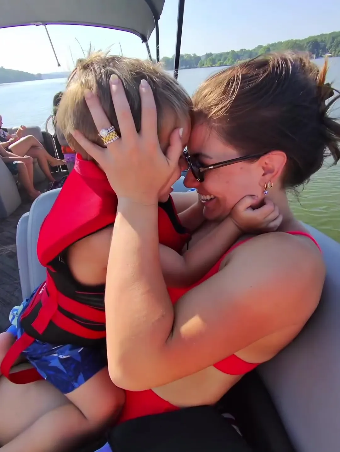 A woman in a red swimsuit and sunglasses embracing a toddler wearing a red life vest on a boat.