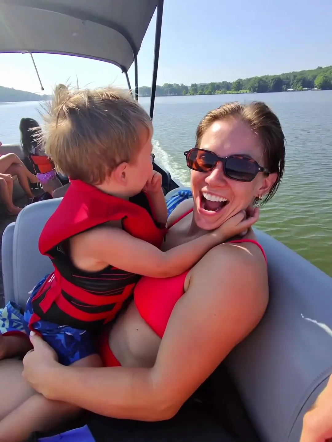 A woman in sunglasses and a red bikini smiles widely as a young child in a life vest hugs her while on a boat.