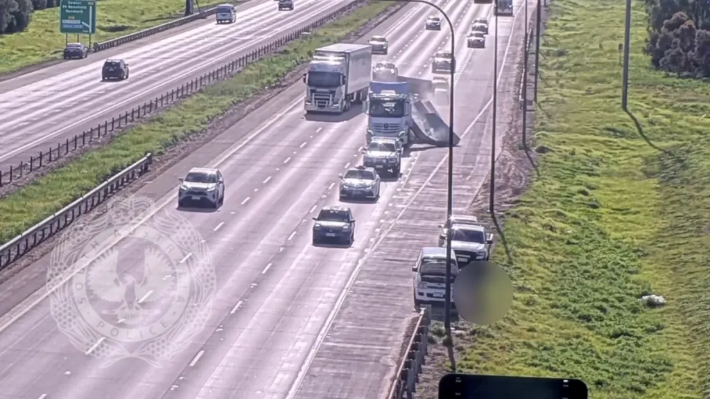 An overhead view of a truck on a highway with concrete slabs falling off the back, causing dust and debris, with cars parked on the shoulder.