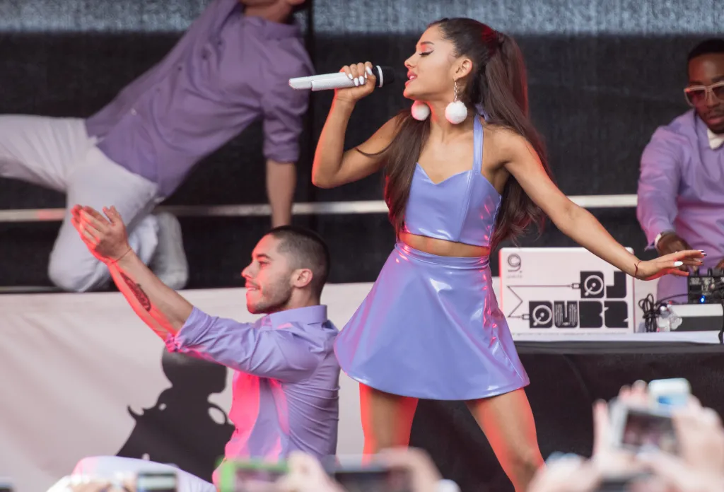Ariana Grande performing on stage, wearing a lavender two-piece outfit, holding a microphone.