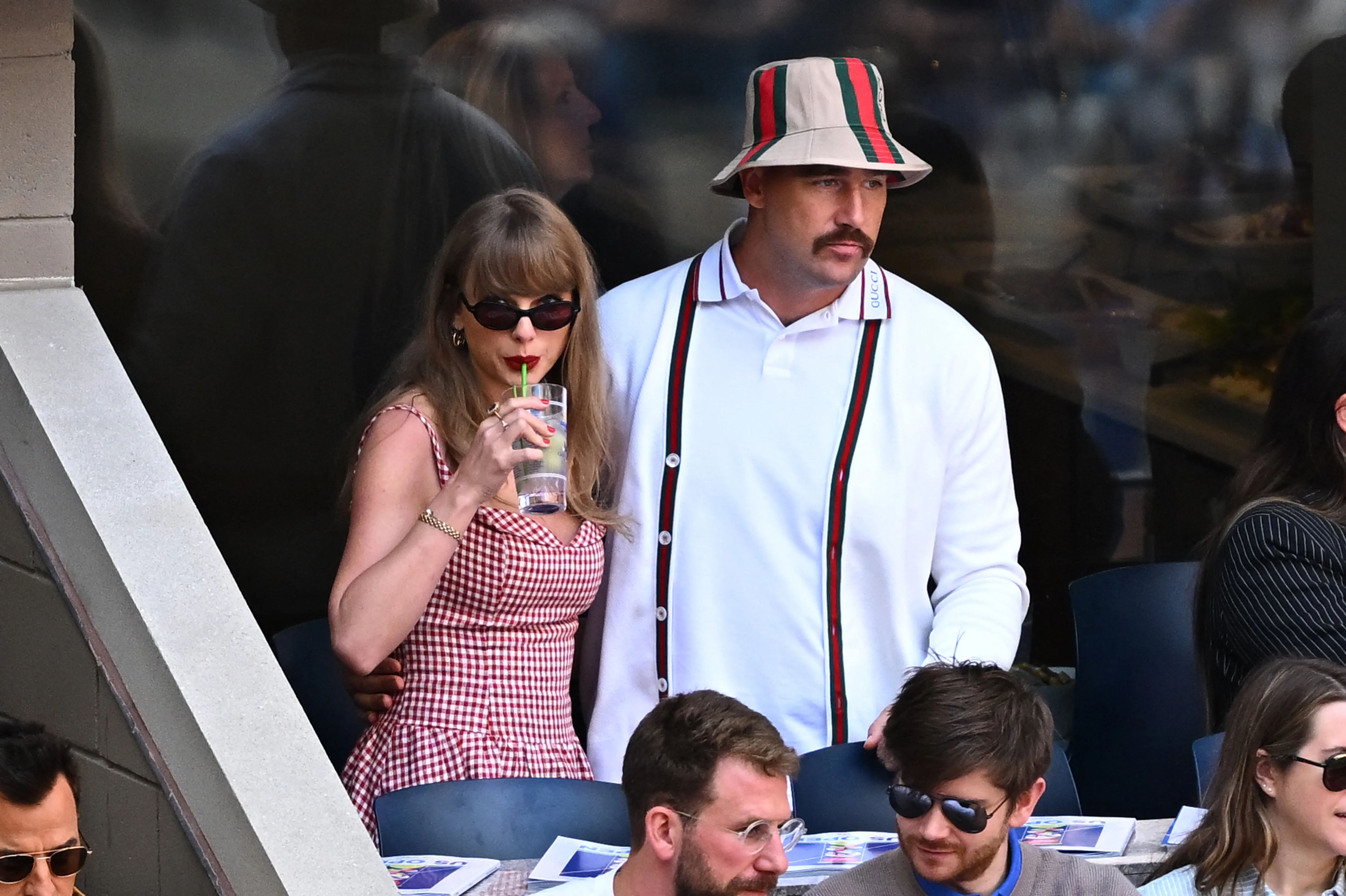 Taylor Swift in a red and white checkered dress and sunglasses, drinking from a glass, with Travis Kelce in a white shirt, striped suspenders, and a striped bucket hat.