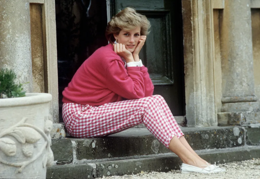Diana, Princess of Wales, in a pink sweater and red and white checkered pants, sits on a step at Highgrove House.