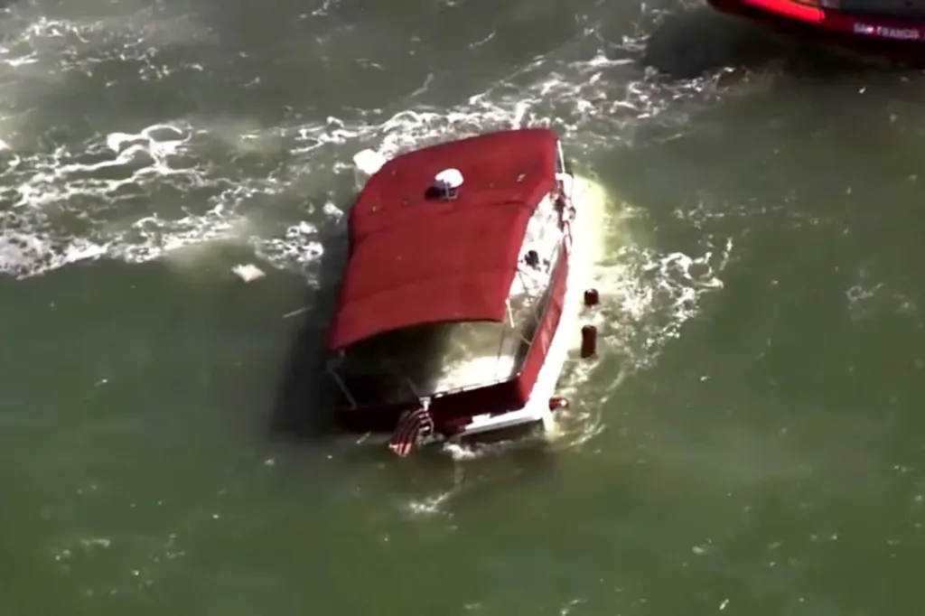 Red and white boat partially submerged in choppy green water, with a Coast Guard boat nearby.