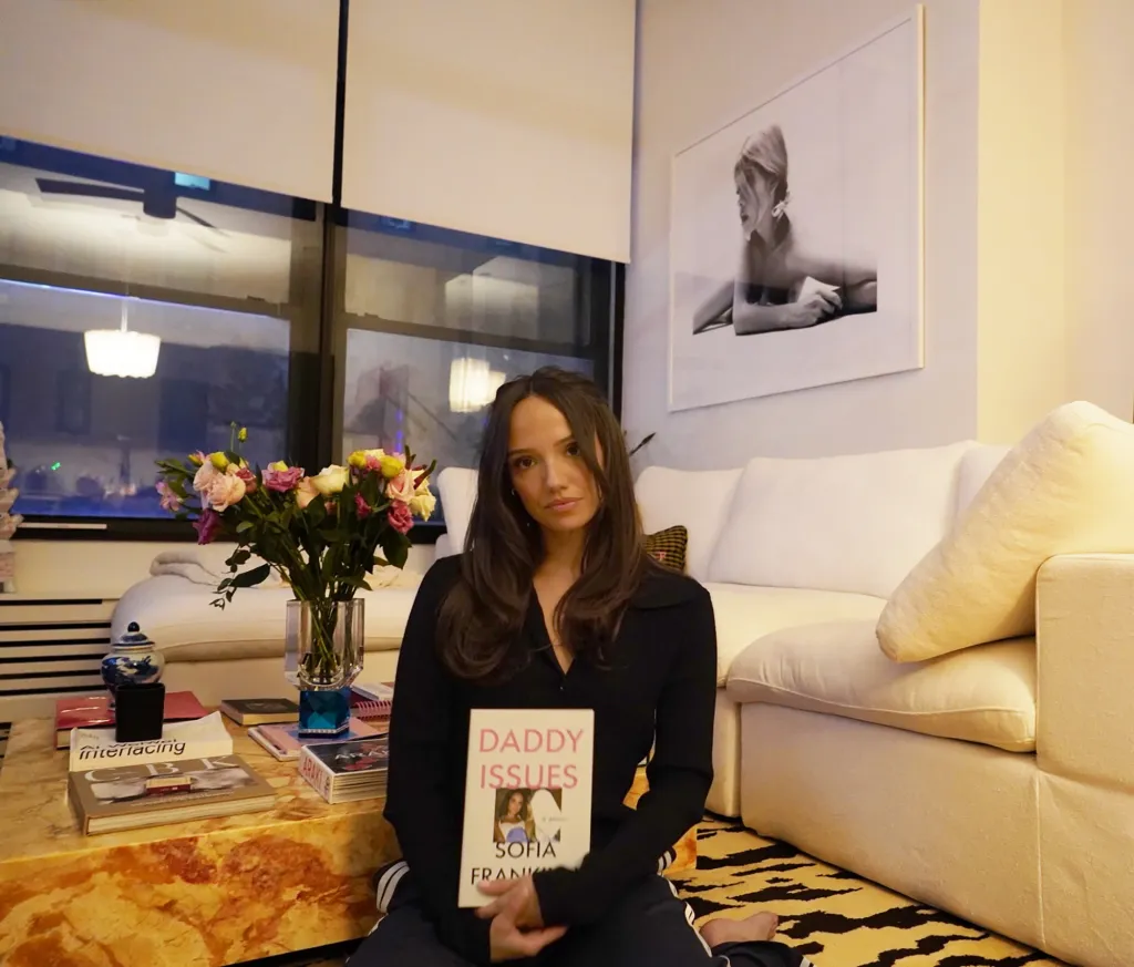 Sofia Franklyn, in a black top and black sweatpants, holding her book "Daddy Issues" in a living room.