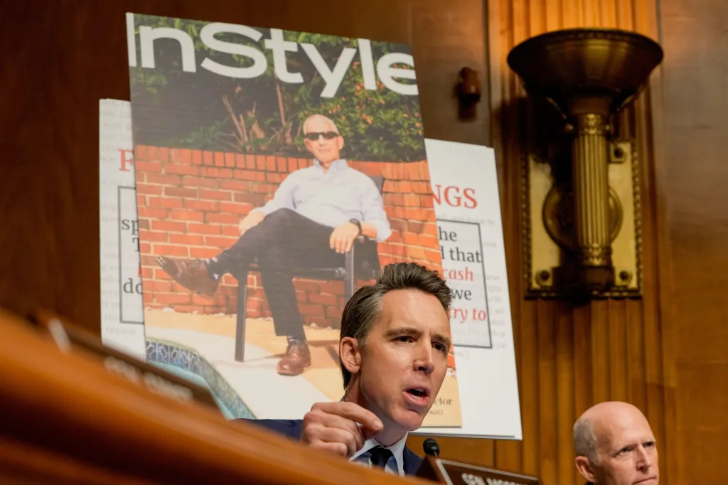 Senator Josh Hawley speaking, with a poster board behind him displaying a magazine cover of Anthony Fauci in a chair with the title "InStyle".
