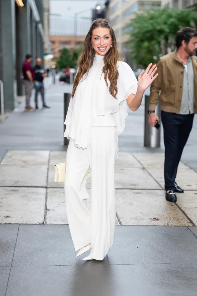 Stassi Schroeder in a white outfit, waving to the camera.
