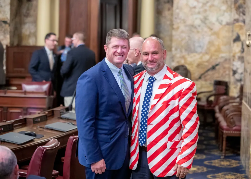 State Rep. Eric Davanzo in a red and white striped jacket and blue tie with white stars, standing next to another man in a blue suit and striped tie.