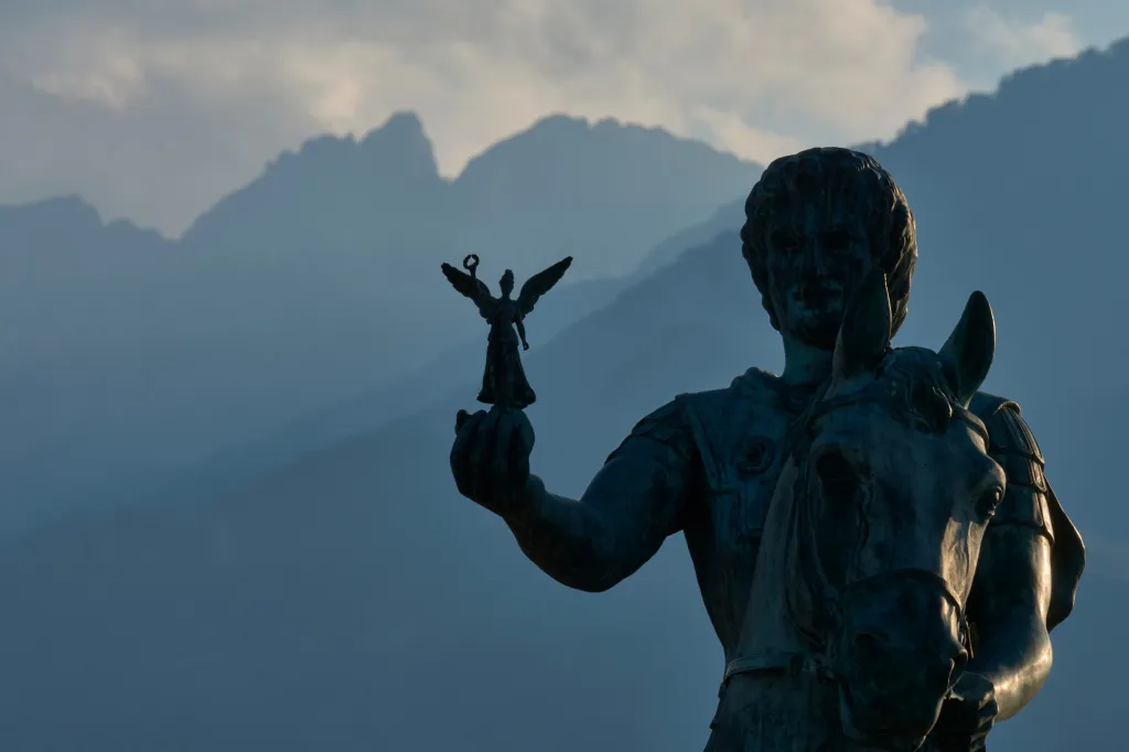 A statue of Alexander the Great on horseback, holding a smaller statue of Nike, with Mount Olympus in the background.