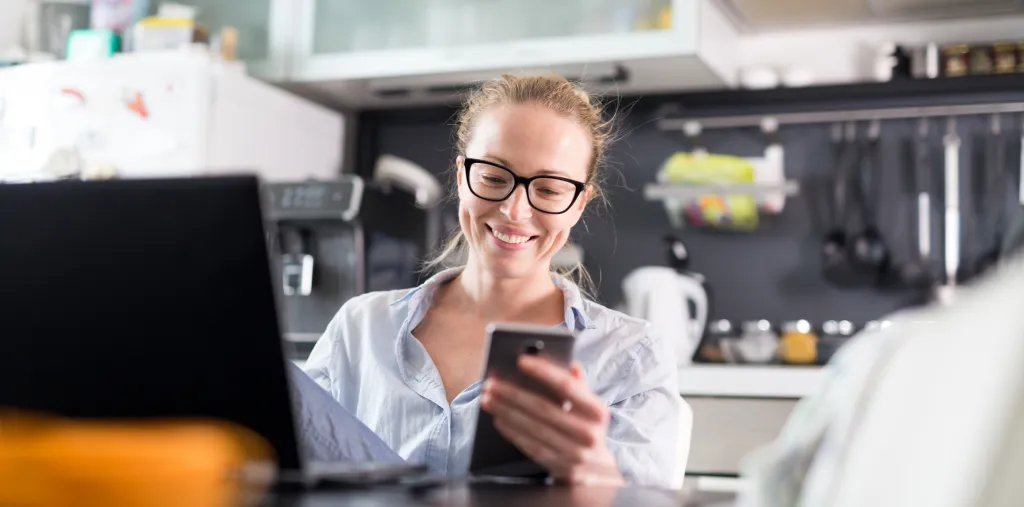 A woman in casual clothing working remotely from her kitchen dining table, video chatting on her smartphone and laptop.