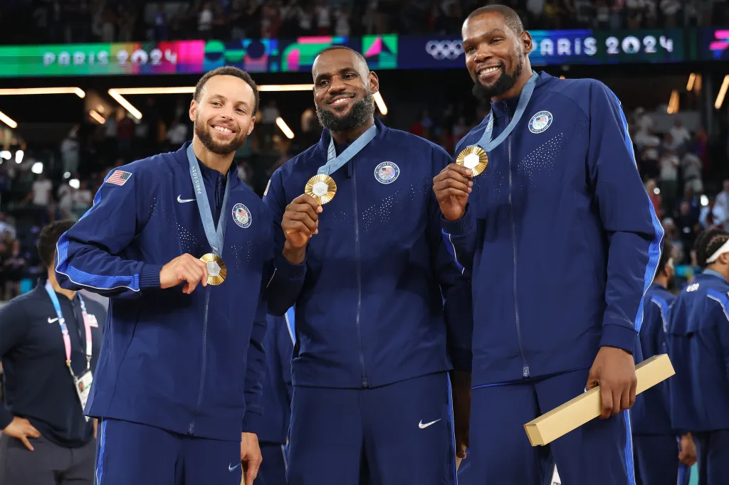 Stephen Curry, LeBron James, and Kevin Durant smiling while wearing gold medals at the Paris 2024 Olympics.