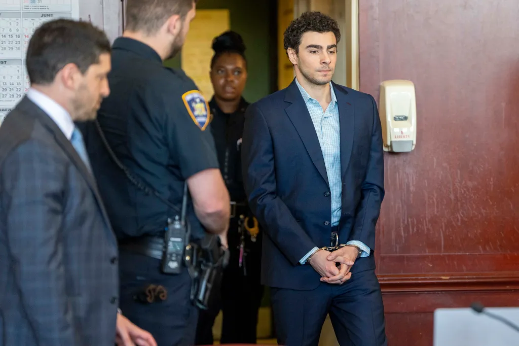 Luigi Mangione, handcuffed in a suit, stands next to two officers and a man in a suit.