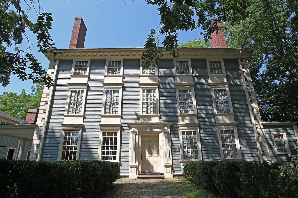 Royall House and Slave Quarters with gray siding, white trim around windows, and a white front door, surrounded by trees and bushes.