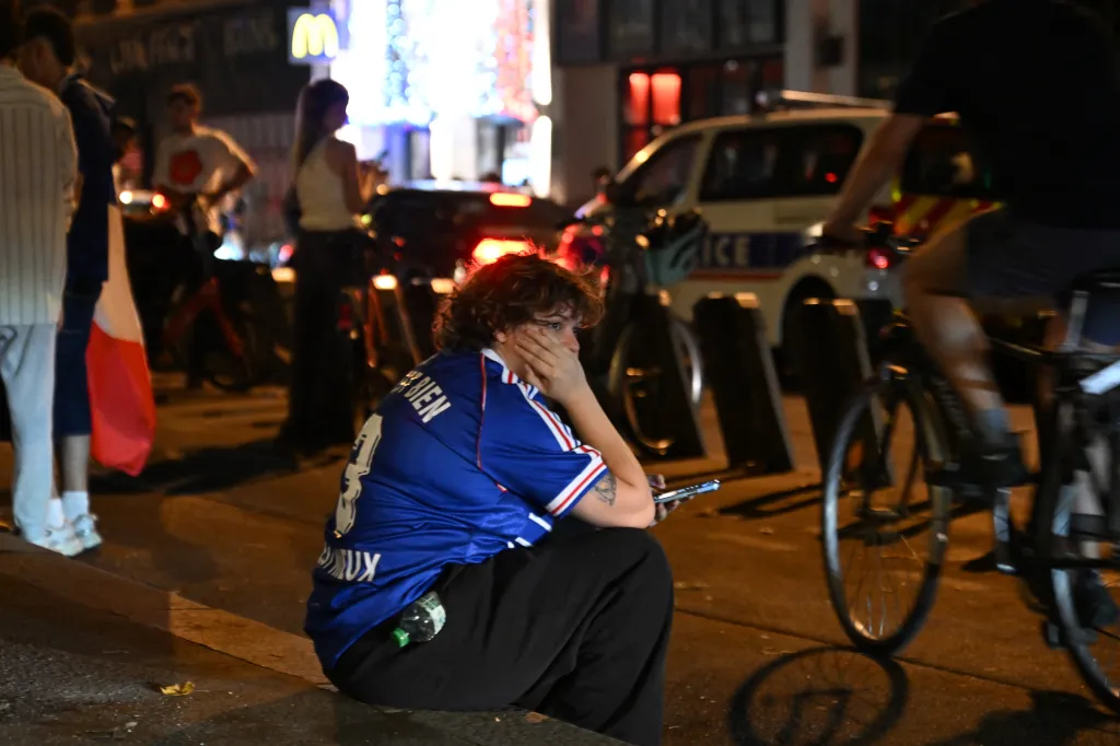 A dejected France football fan sits on the curb, holding a phone, after the World Cup semi-final loss.