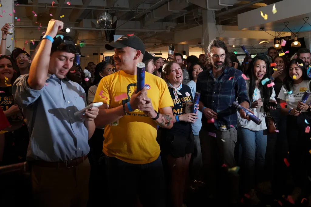 Supporters at a primary election night watch party celebrating Melat Kiros's lead, with confetti falling and people holding noisemakers and drinks.