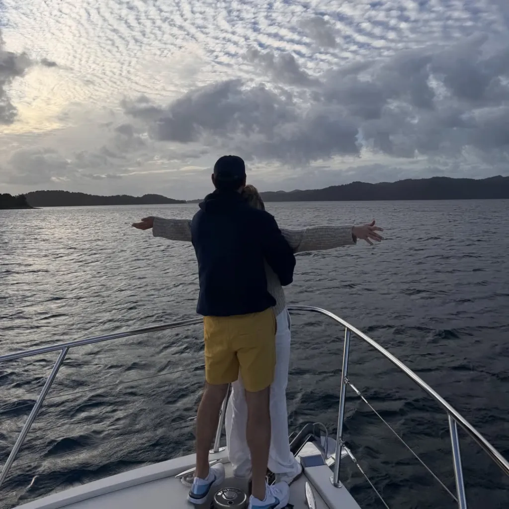 Sydney Sweeney and Scooter Braun embracing on a boat with arms outstretched, looking out over the water.