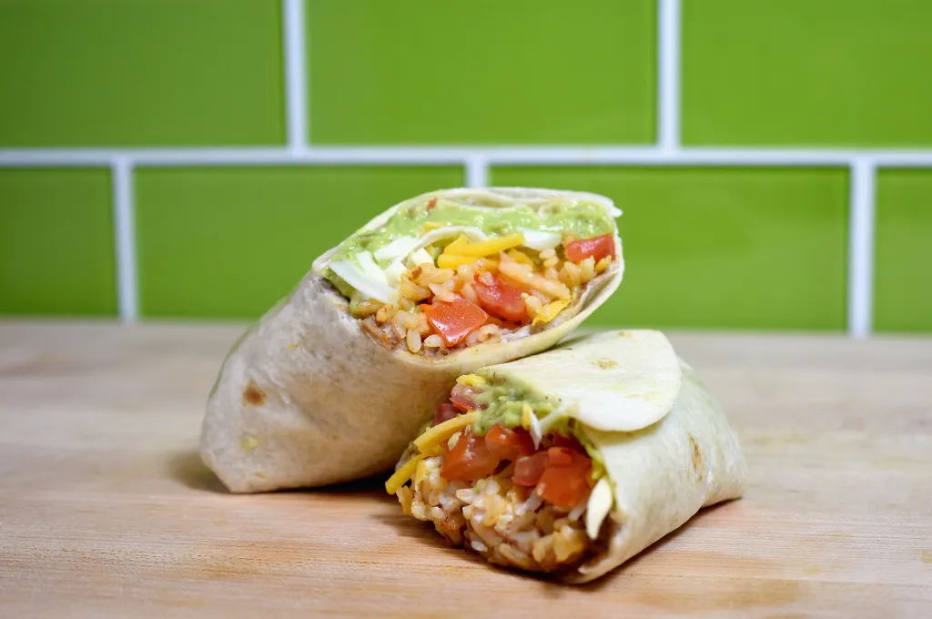 Two halves of a Taco Bell 7-Layer Burrito, exposing its ingredients, sit on a wooden surface with a green tiled wall in the background.