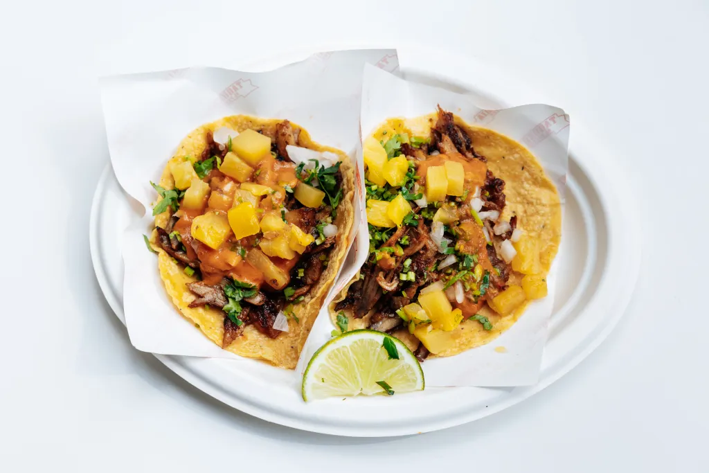Two al pastor tacos with pineapple, cilantro, and onions, served on a white plate with a lime wedge.