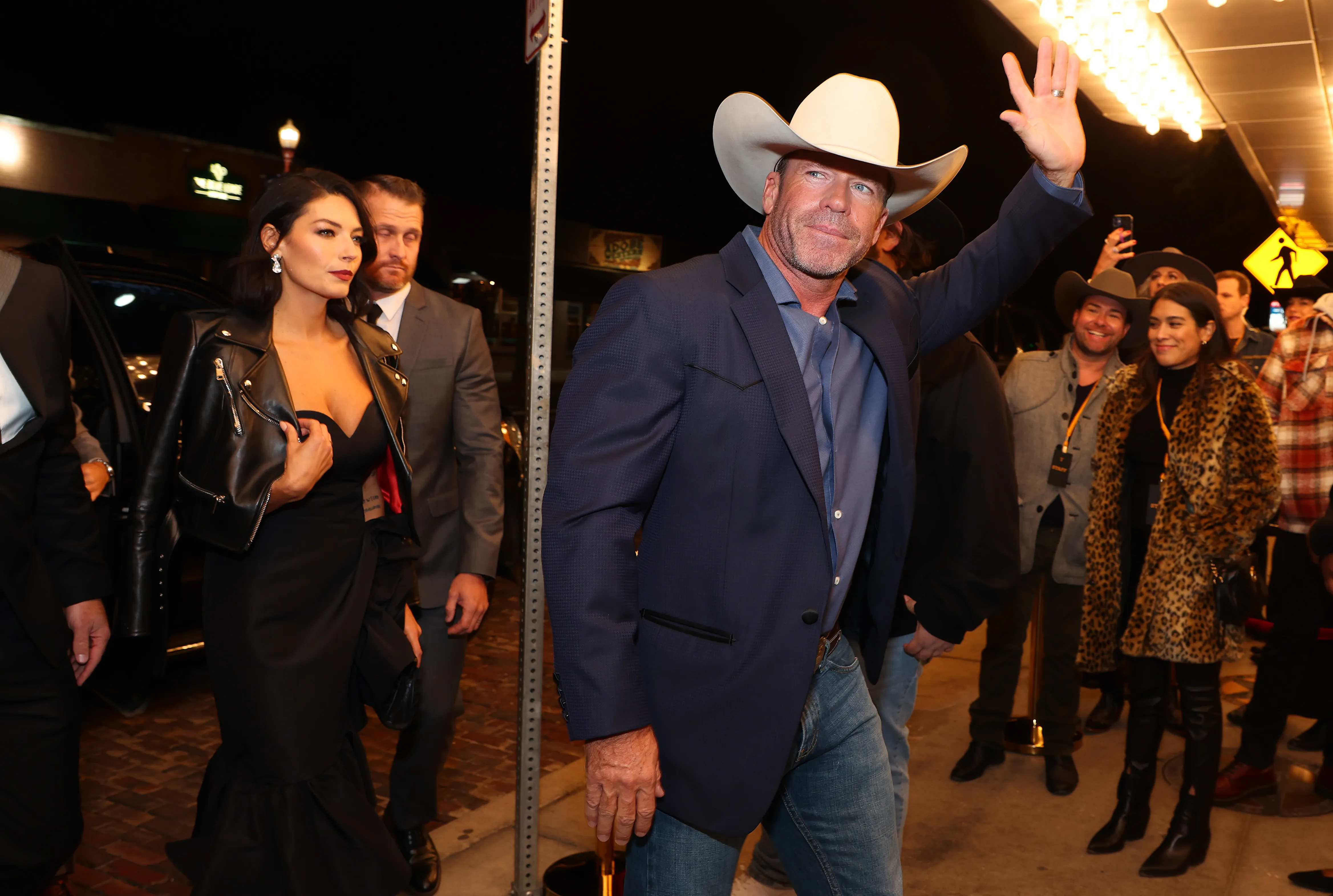 Taylor Sheridan, in a cowboy hat and blazer, waves to the crowd while Nicole Sheridan poses next to him in a black dress and leather jacket.