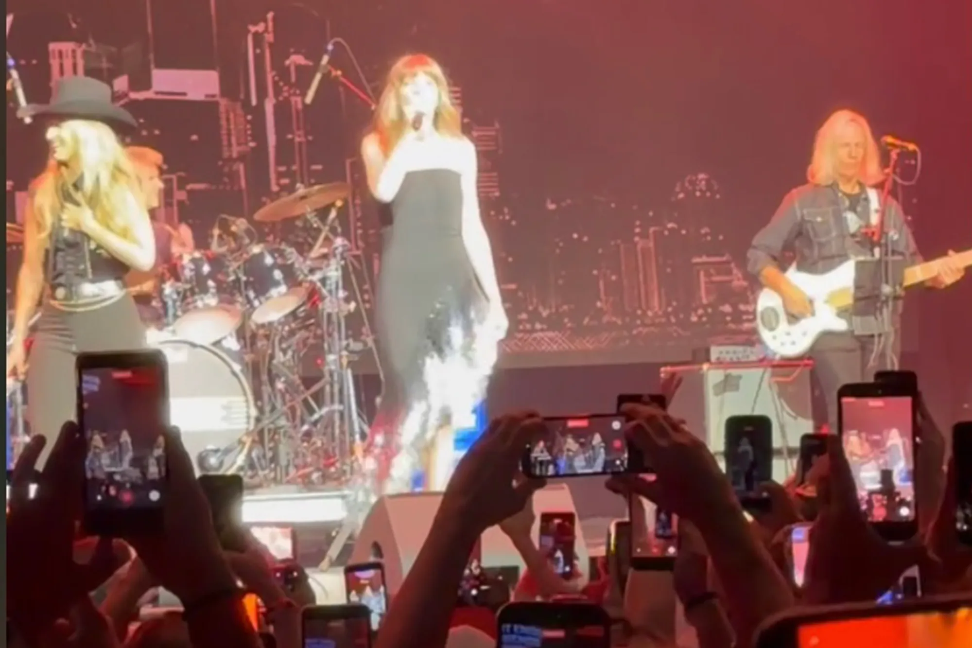 Taylor Swift performing on stage in a black dress, with a band and audience recording on phones.