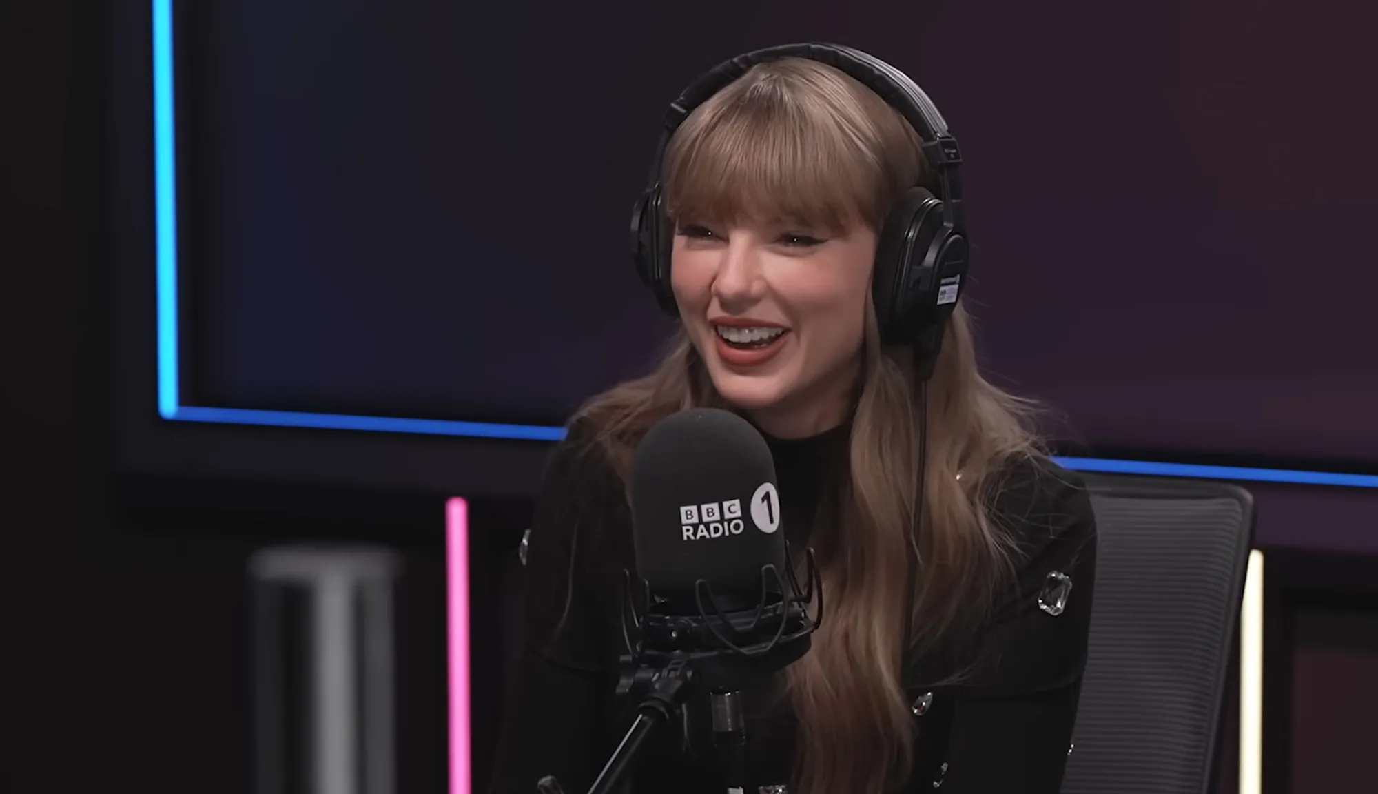Taylor Swift laughing while wearing headphones and speaking into a BBC Radio 1 microphone.