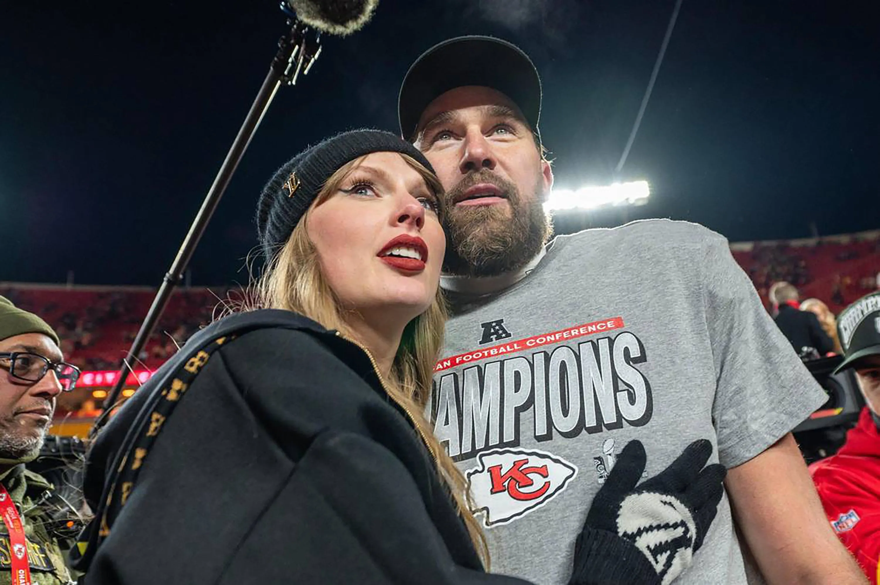 Taylor Swift in a black beanie and Travis Kelce in an "AFC Champions" t-shirt looking up.