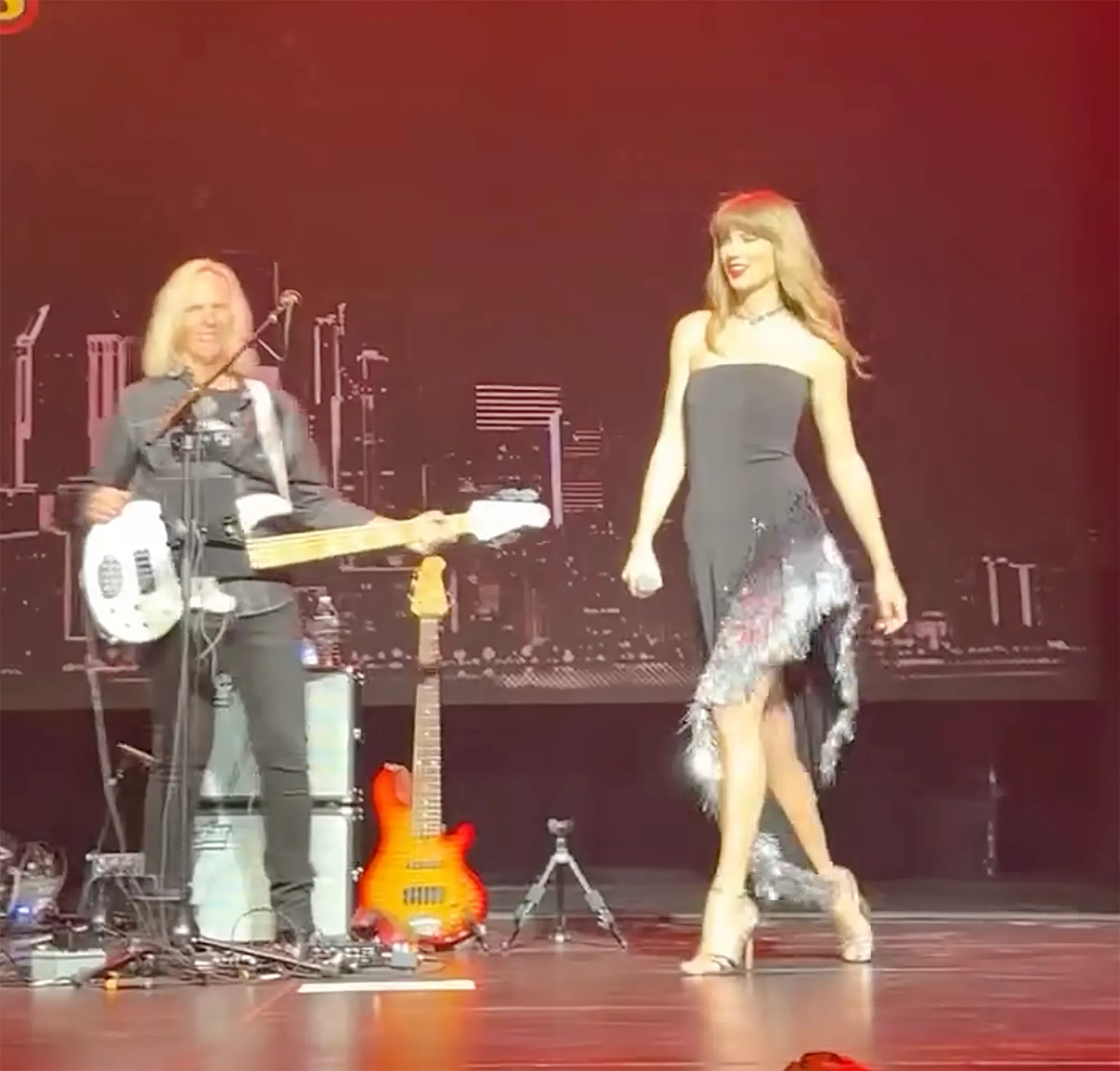Taylor Swift performing on stage in a black dress with a musician in the background.