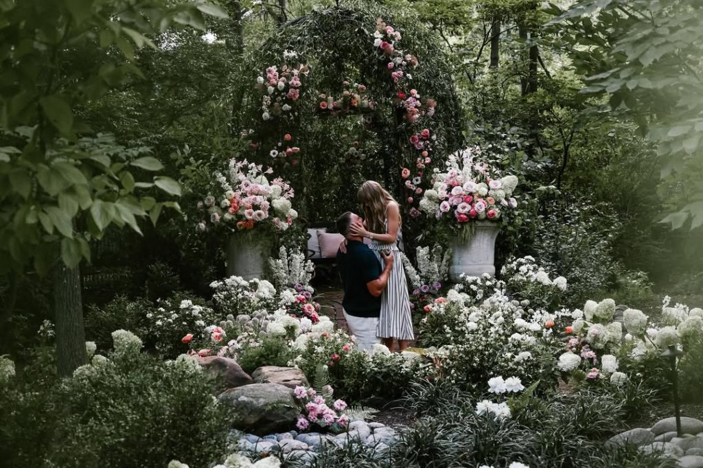 Travis Kelce on one knee proposing to Taylor Swift in a lush garden filled with white and pink flowers.