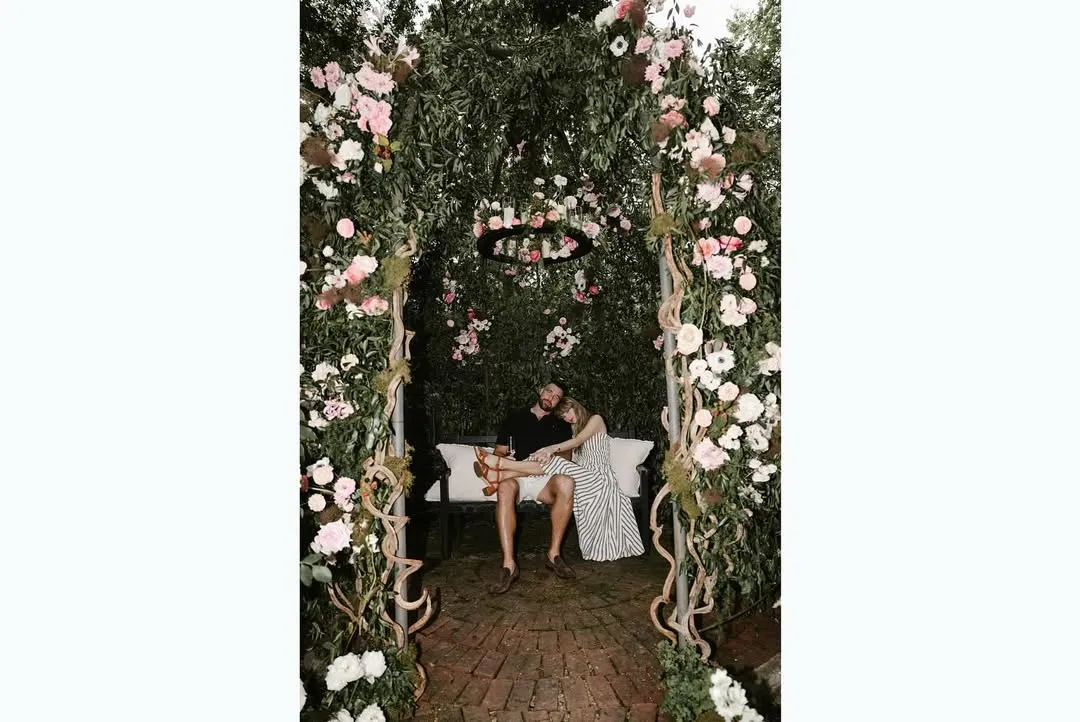 Taylor Swift and Travis Kelce sitting on a bench beneath a flower-laden archway.