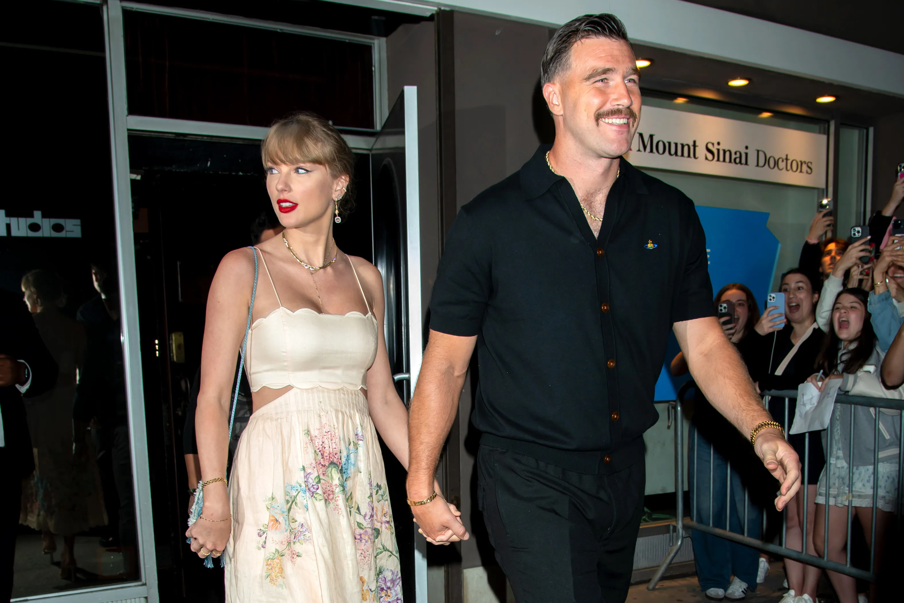 Taylor Swift in a white floral dress and Travis Kelce in a black shirt holding hands, surrounded by people holding up phones.