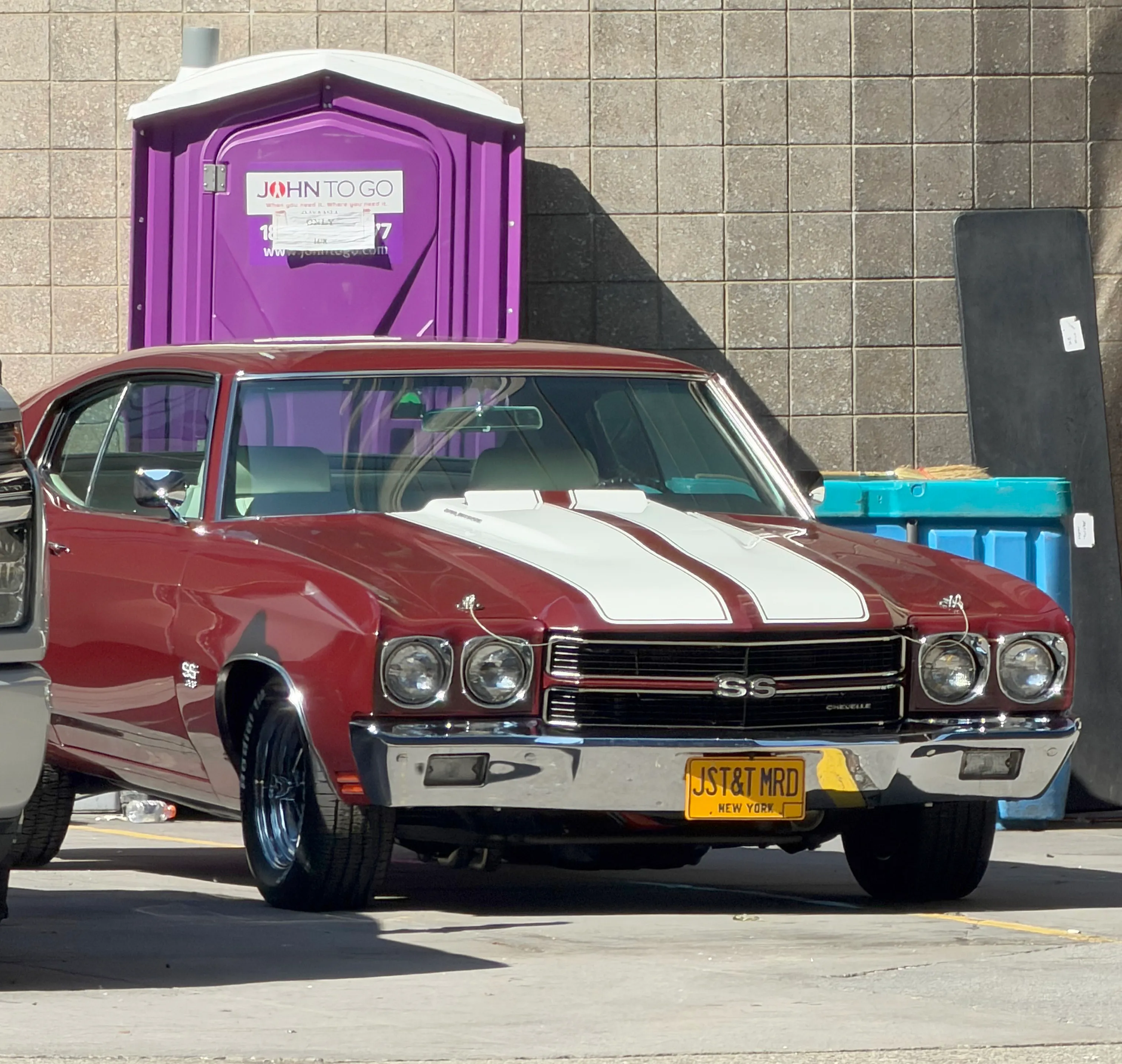 A maroon Chevrolet Chevelle with white racing stripes and a license plate reading "JST&T MRD" is parked in front of a purple portable toilet.