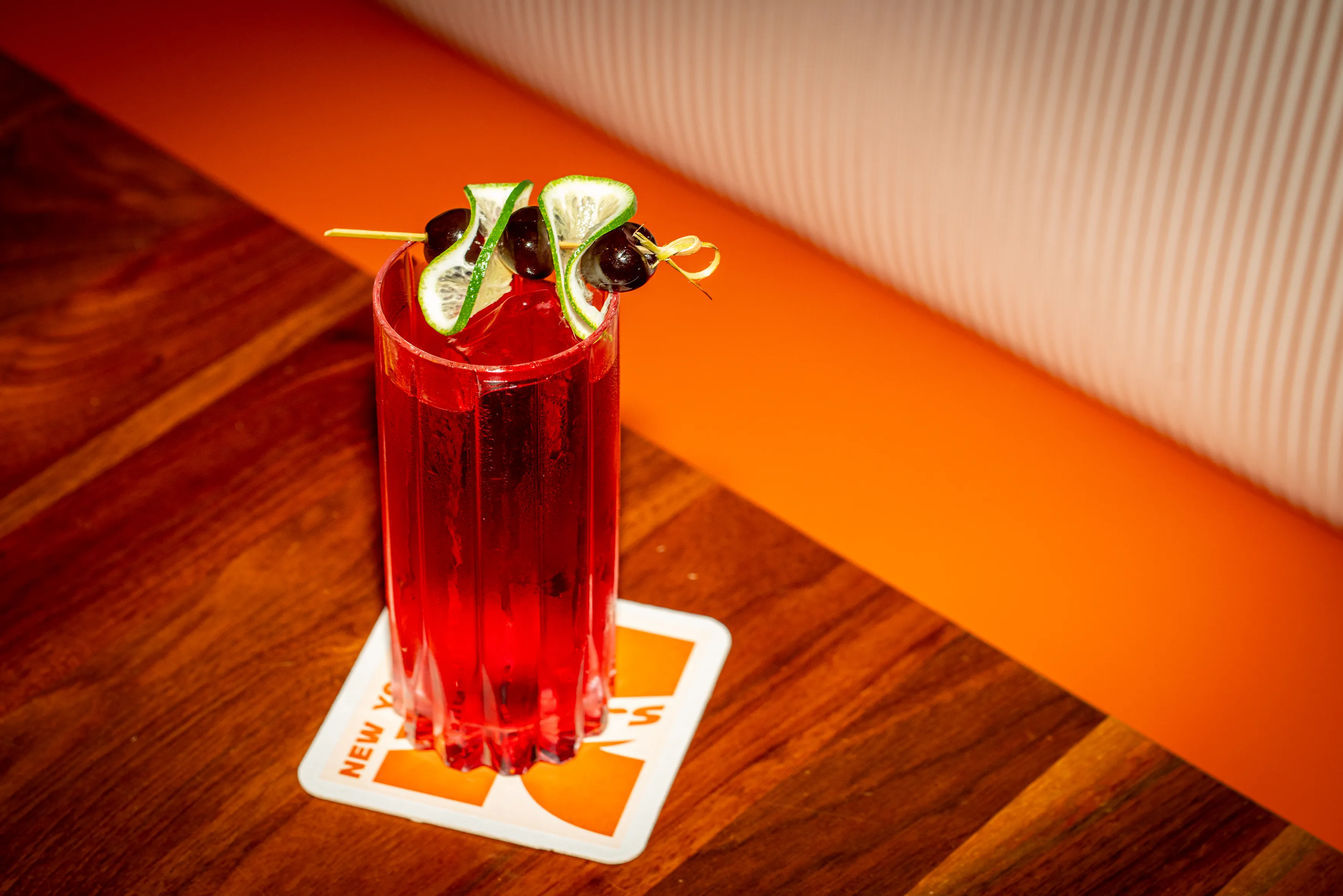 A red cocktail in a tall glass with a skewer of limes and cherries on a branded coaster.