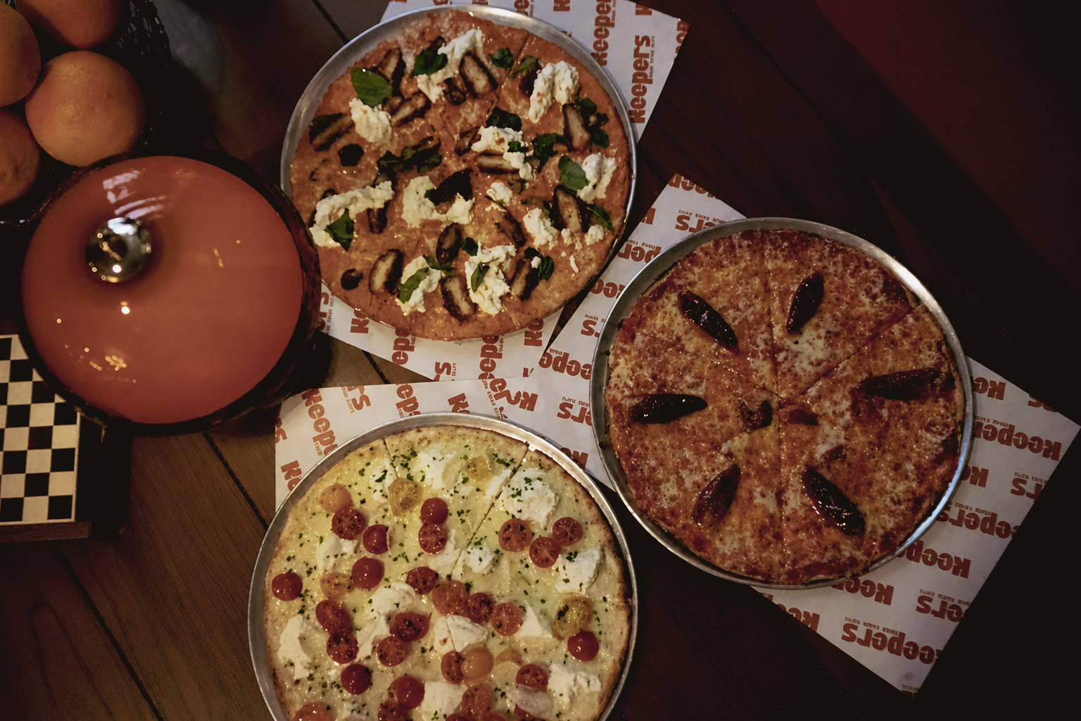 Three pizzas, one with red and yellow cherry tomatoes and white cheese, another with green leaves and white cheese, and a third with dark toppings, arranged on a table with an orange pot and oranges.
