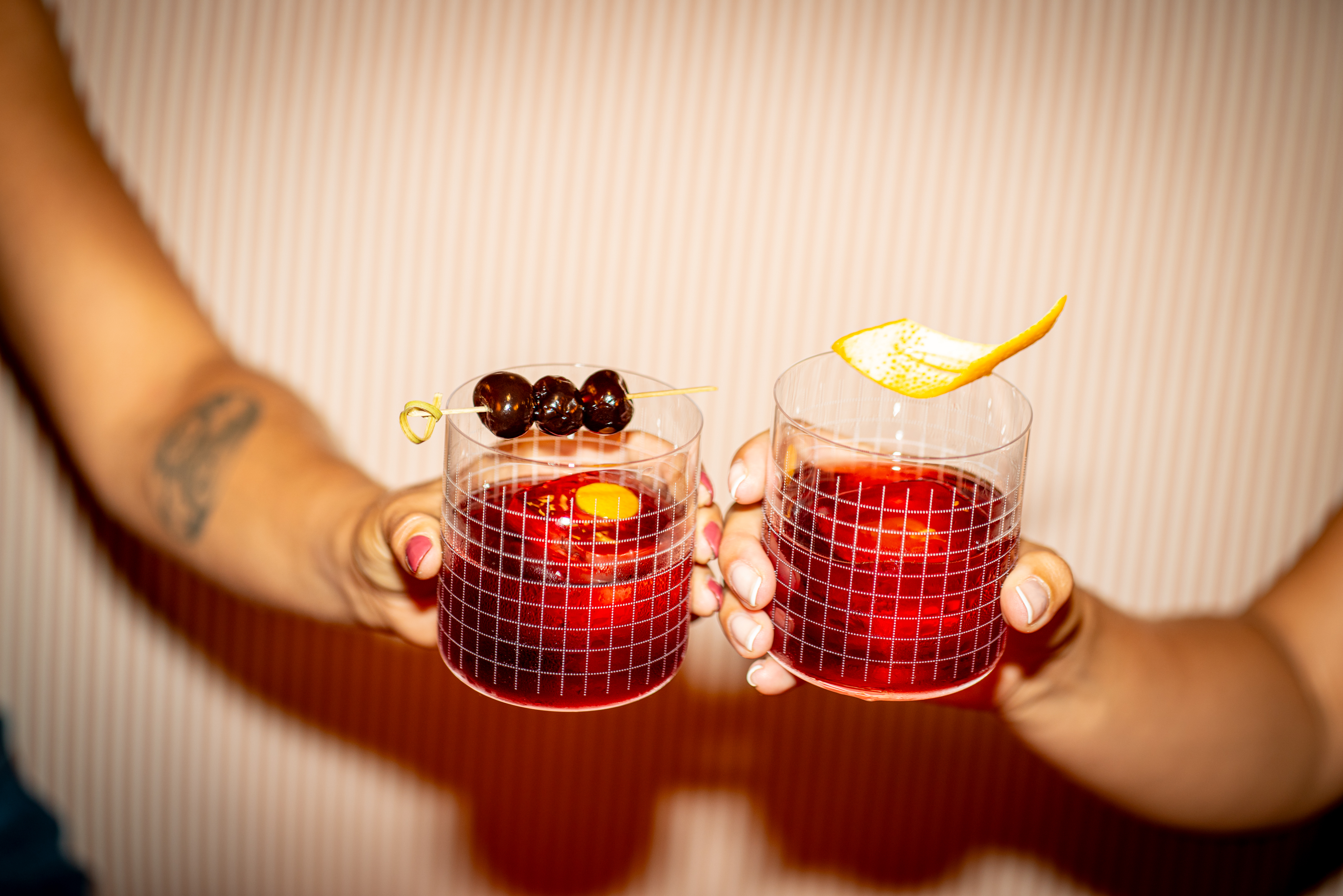 Two hands holding out glasses of dark red cocktails, one garnished with cherries, the other with an orange peel.