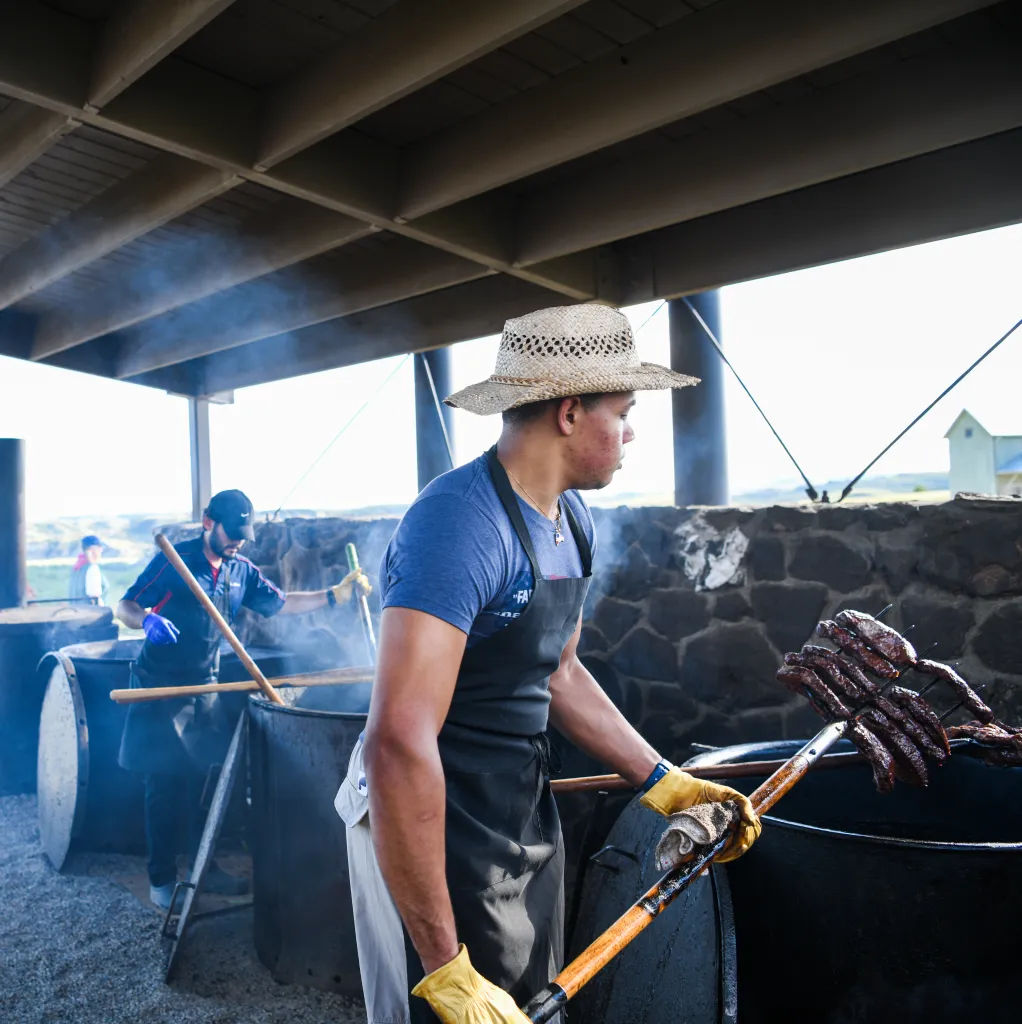 Two men cooking "Pitchfork Fondue" by placing meat on pitchforks into large vats.