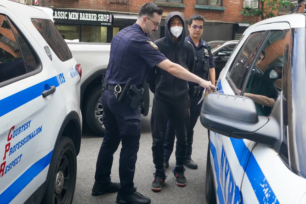 A police officer leading a young man in a black hoodie and white mask towards a police car, with another officer standing nearby.