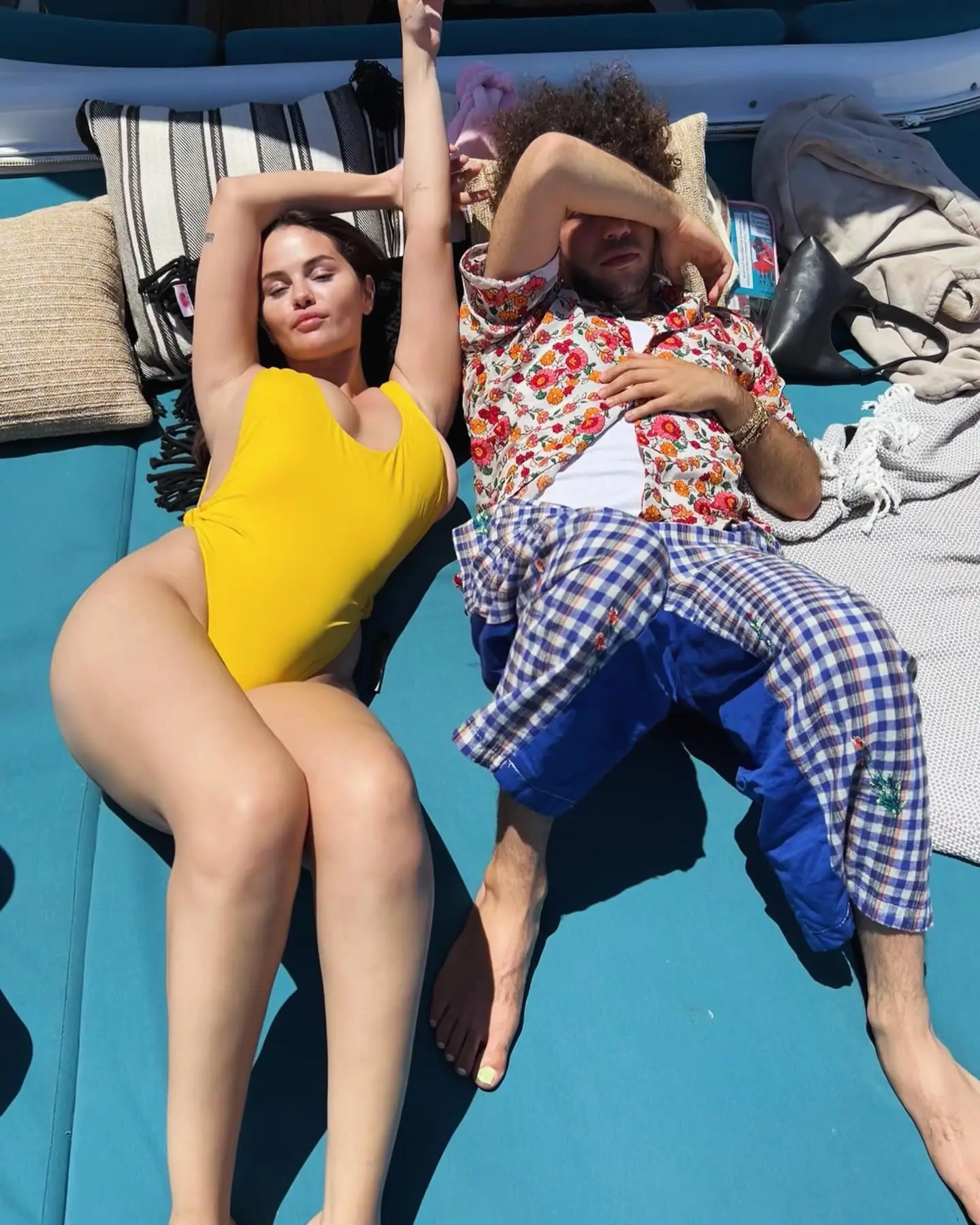 Selena Gomez in a yellow swimsuit and Benny Blanco in a floral shirt and plaid pants relaxing on a boat.