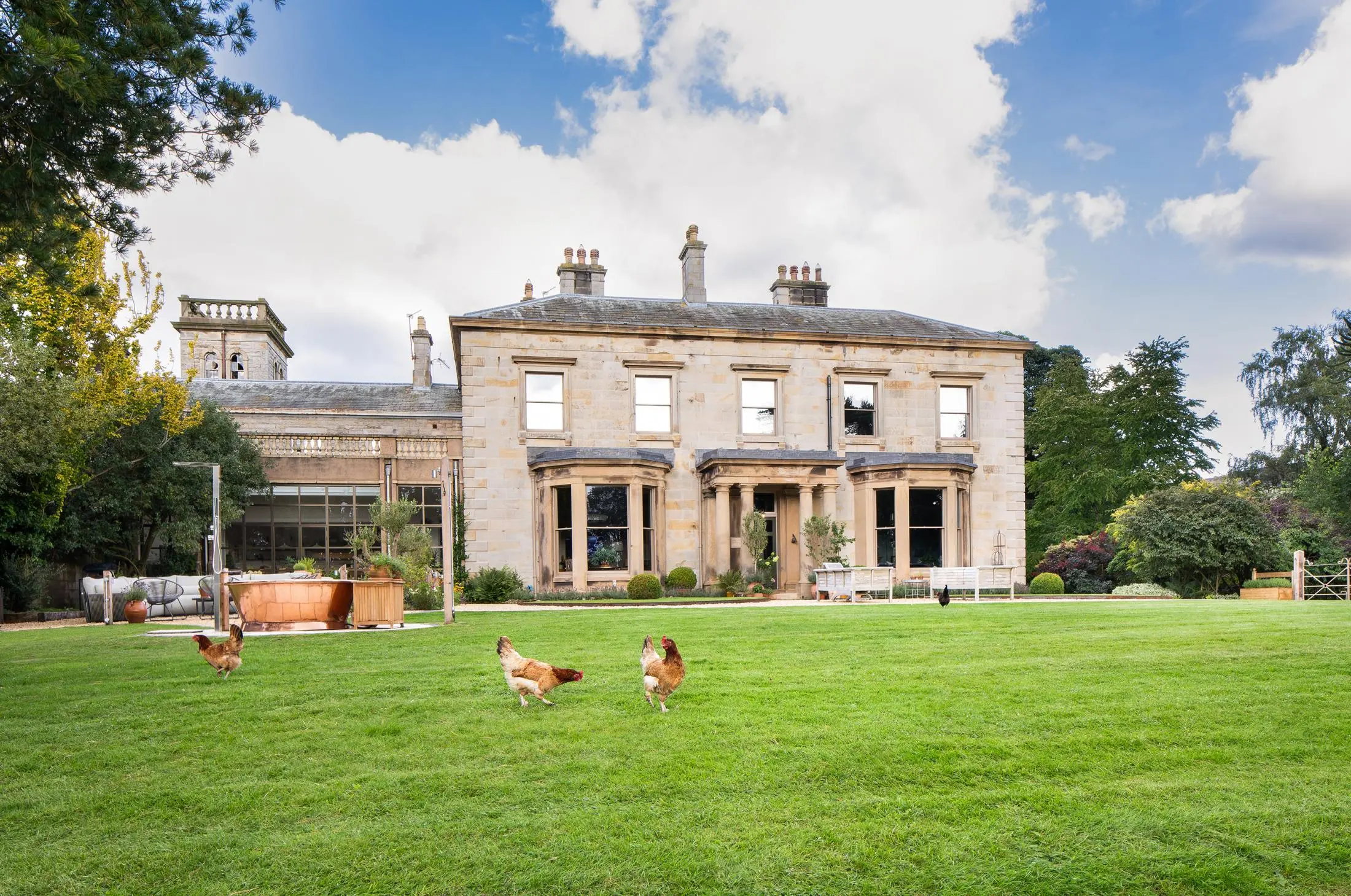 The Country House Cumbria, a large stone building with a green lawn and chickens in the foreground.