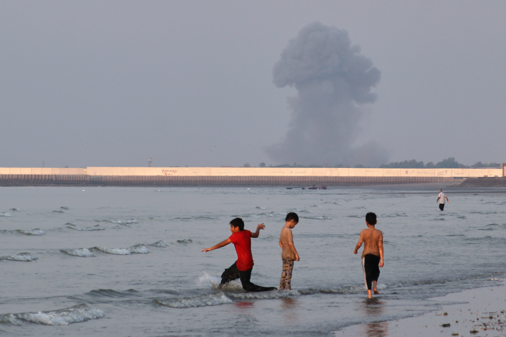 Three boys play in the shallow waters of the Strait of Hormuz as a plume of smoke rises from an explosion in the background, off Bandar Abbas, Iran.